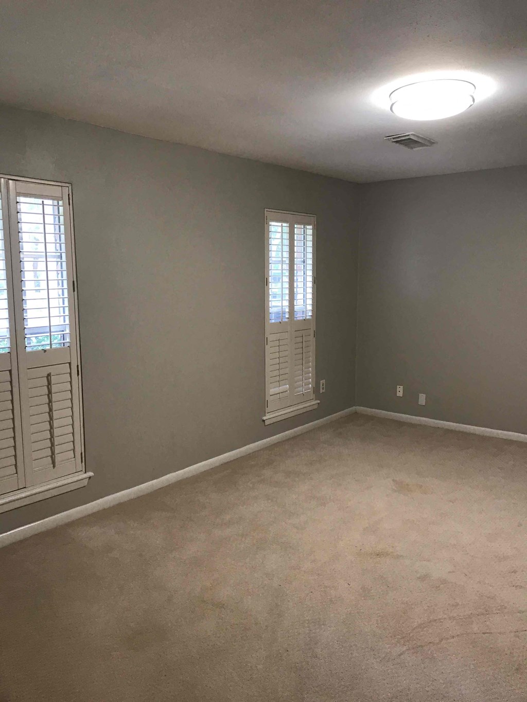 A room with a carpeted floor and two windows with shutters.
