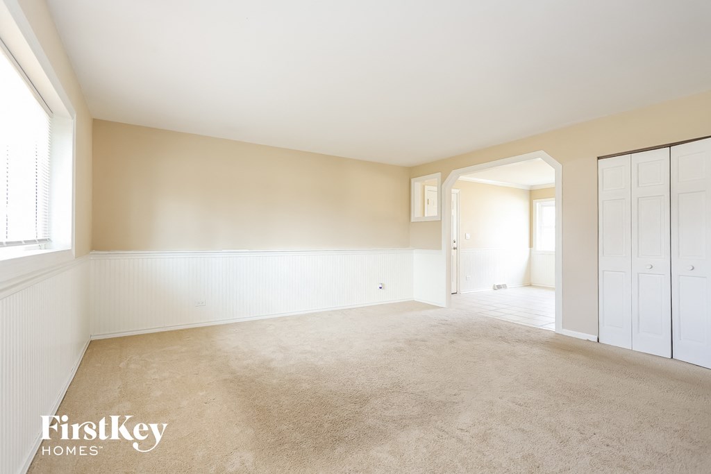 A spacious room with beige carpeting and white walls, with a window on the left and a door on the right.