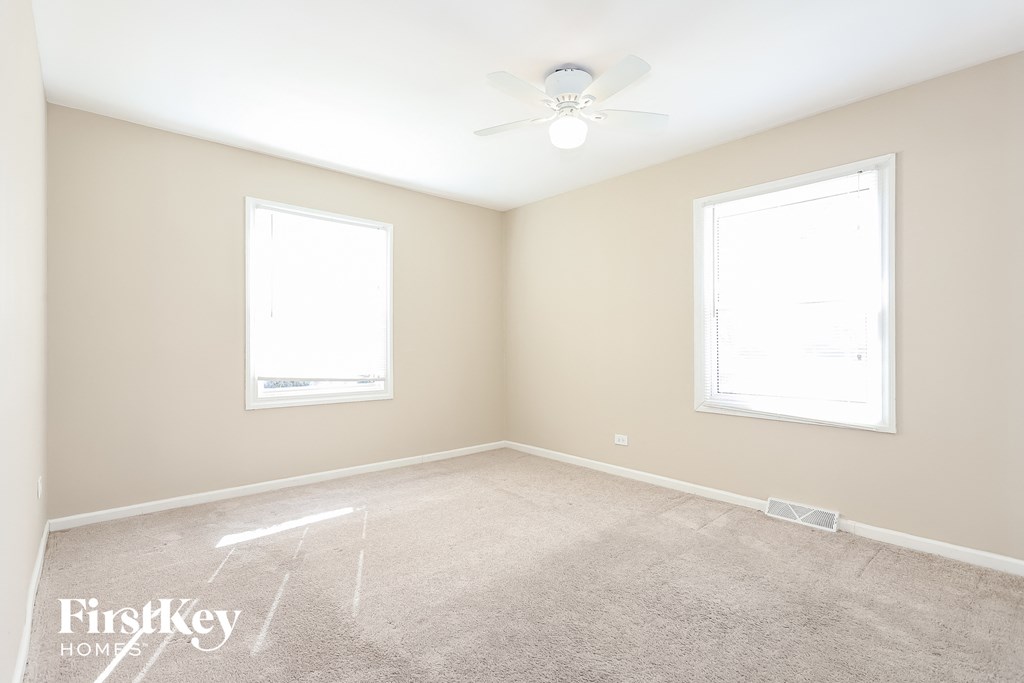 A room with a carpet, a ceiling fan, and two windows.