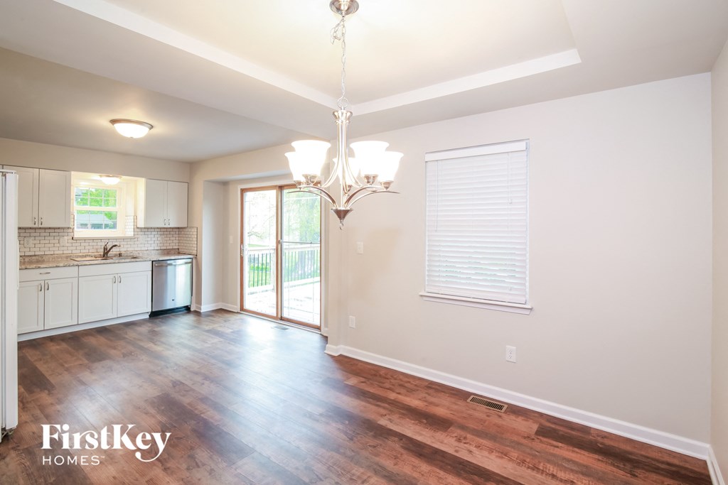 A well-lit, empty room with a chandelier and a view of a kitchen area.