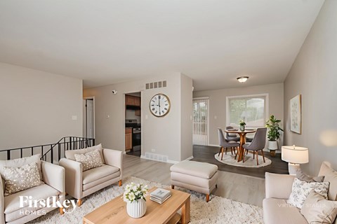 A living room with a clock on the wall and a dining table with chairs.