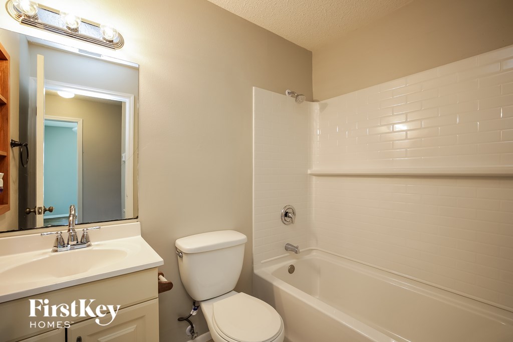 A white toilet sits in a bathroom with a white tub and a mirror above a sink.