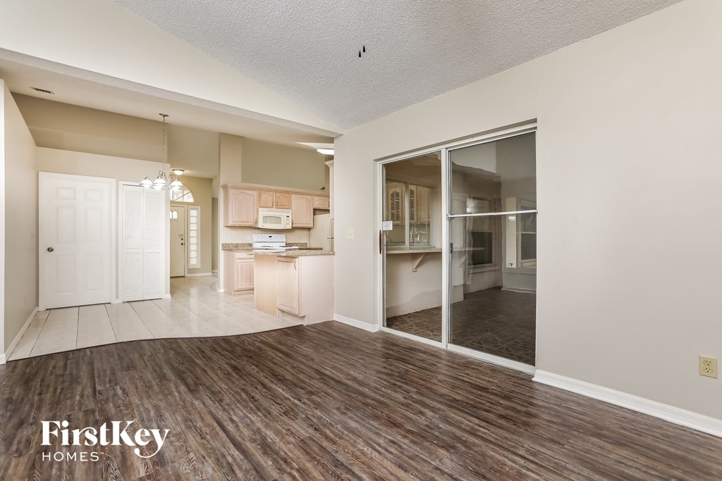 A spacious living room with wood flooring and a kitchen visible through the glass doors.