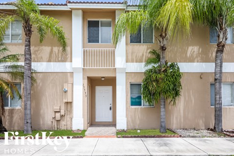 A beige two-story apartment building with a balcony and palm trees in front.