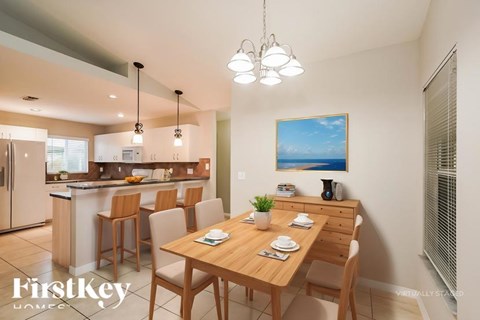 A dining room with a wooden table and chairs, a painting on the wall, and a kitchen area in the background.