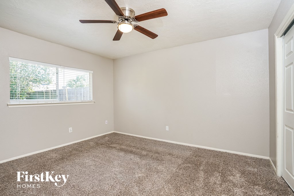 A room with a carpet floor, a ceiling fan, and a window with a view of a fence and trees.