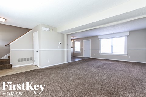 A spacious living room with a carpeted floor and a staircase on the left side.