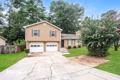 A house with a brown siding and a white garage door is surrounded by a fence and a tree with red flowers.