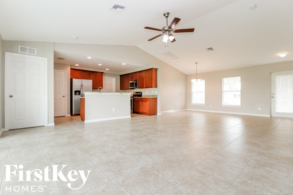 A spacious empty room with a ceiling fan and a kitchen area in the background.
