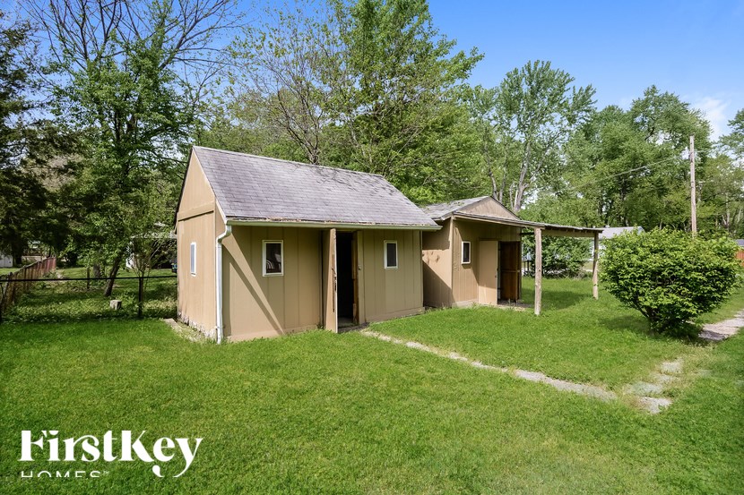 A small house with a brown roof and a white fence is surrounded by green grass and trees.