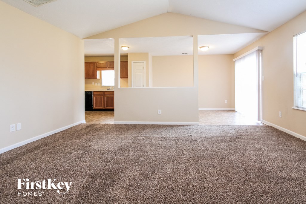 A spacious empty room with a carpeted floor and a kitchen visible through a glass door in the background.