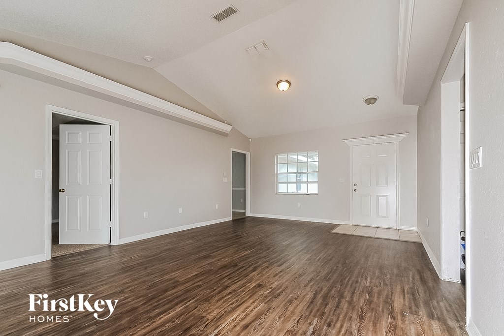 A spacious room with wooden floors and white walls, with a door on the left and a window on the right.