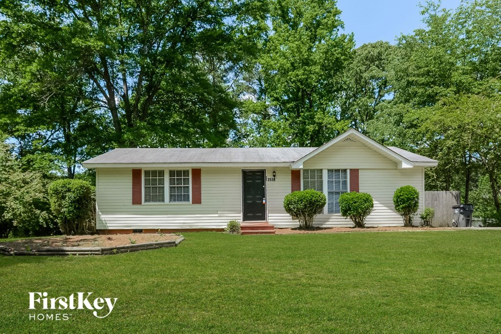 A white house with a black door and red window frames is surrounded by greenery.
