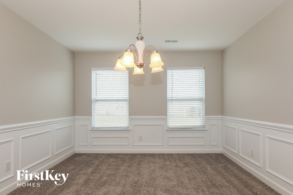 A room with a carpeted floor, two windows with blinds, and a chandelier hanging from the ceiling.