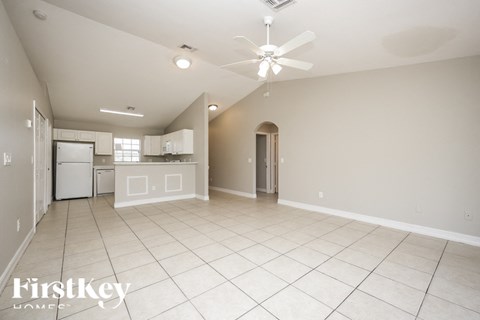 A spacious empty room with a ceiling fan and a kitchen area in the background.