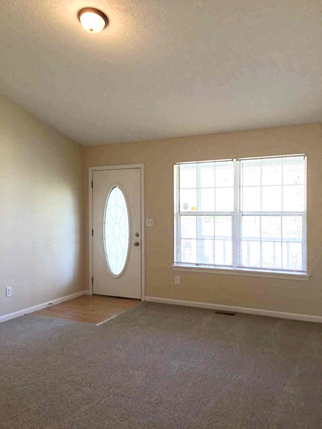 A room with a carpeted floor, a white door with a glass window, and a window with blinds.