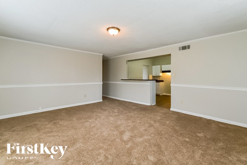 A room with a carpeted floor and a small kitchen area in the background.