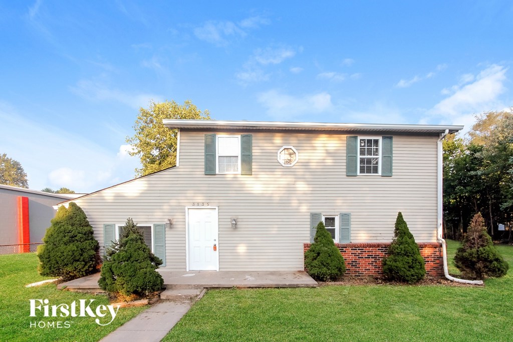 A house with a grey facade and a white door is surrounded by greenery.