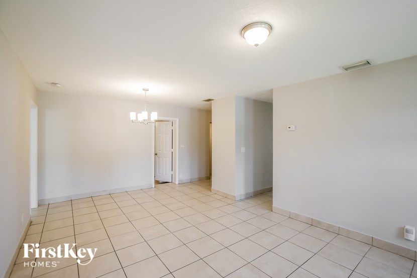 A spacious room with white walls and tile flooring, featuring a chandelier and a doorway leading to another room.