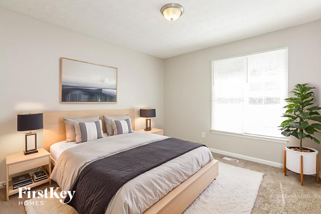 A bedroom with a large bed, a nightstand with a lamp, a framed picture on the wall, and a potted plant on the side table.