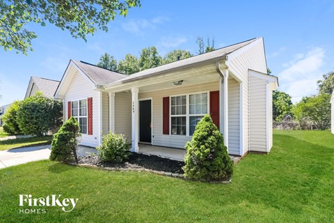 A white house with red shutters and a black door is surrounded by greenery.