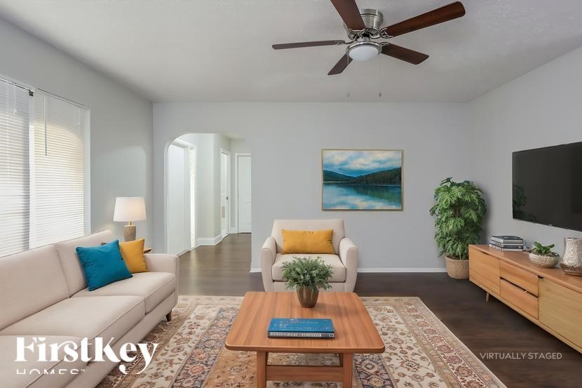 A living room with a couch, a chair, a coffee table, a plant, a painting, a ceiling fan, and a flat screen TV.
