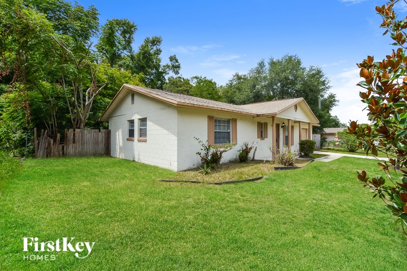 A small white house with a brown roof is surrounded by a green lawn and trees.