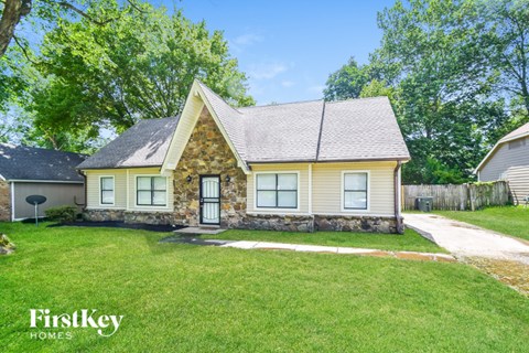 A house with a stone wall and a brown roof is surrounded by a grassy lawn.