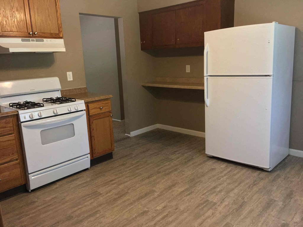 A white refrigerator stands in a kitchen with a white stove and wooden cabinets.