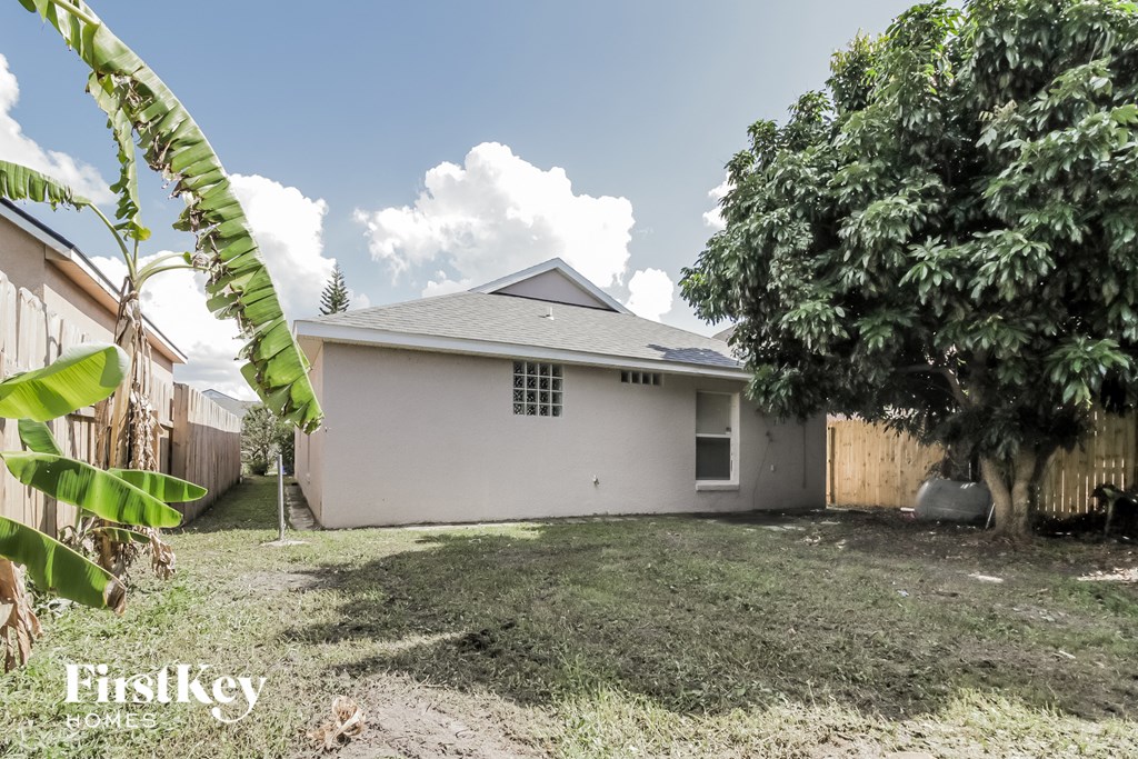 A white house with a grey roof is surrounded by greenery and a fence.