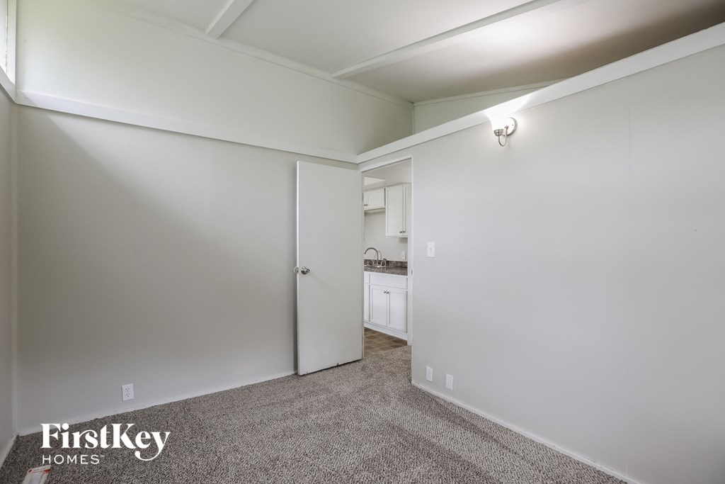 a bedroom with white walls and carpet and a door to a kitchen