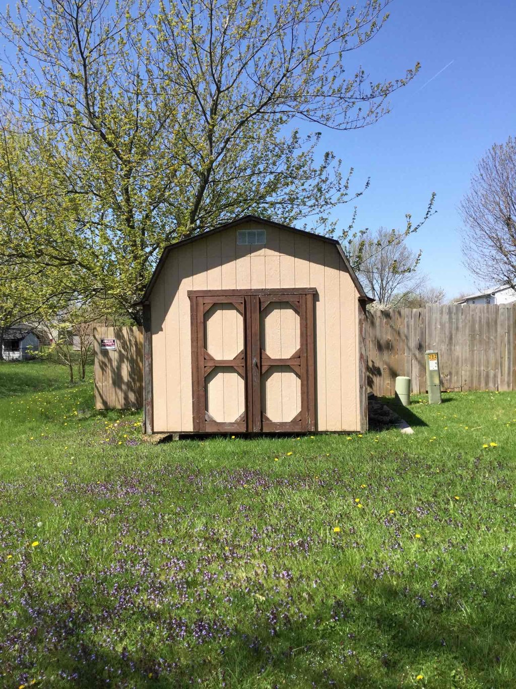 A small wooden shed with a brown door is situated in a grassy area with purple flowers.