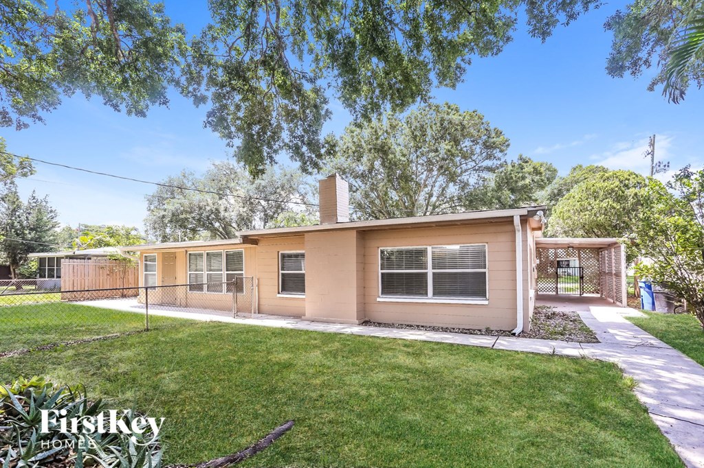 A beige house with a brown roof and a white fence is surrounded by green grass and trees.