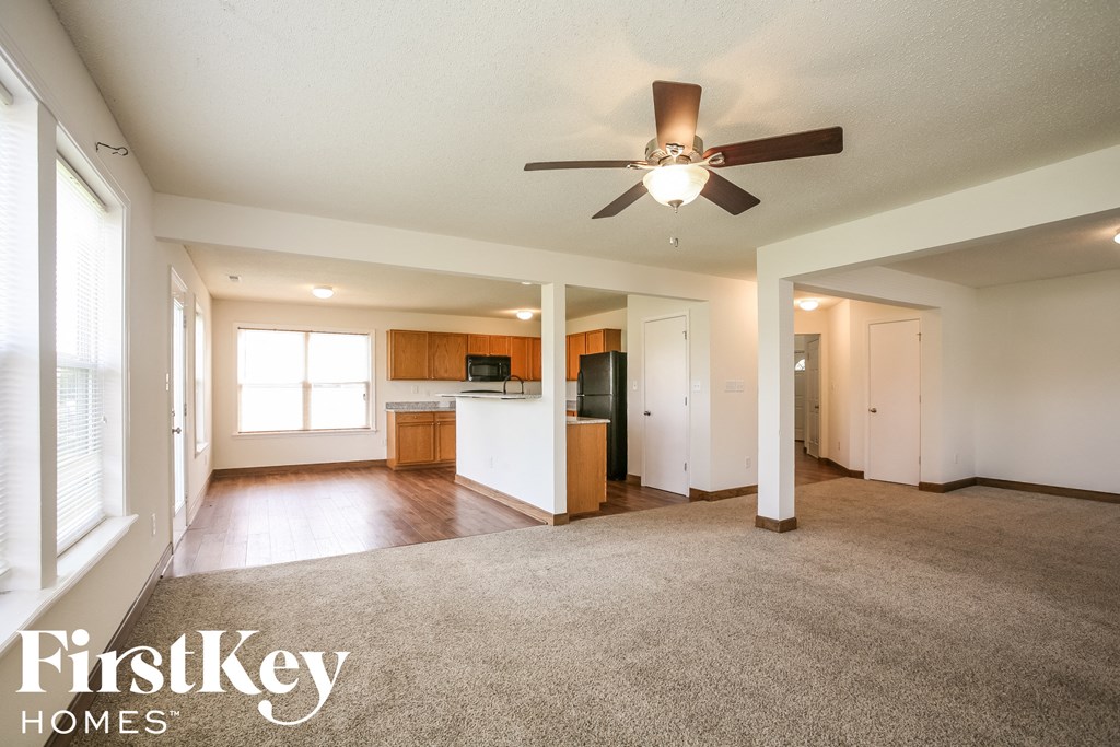 A spacious living room with a ceiling fan and a kitchen area in the background.