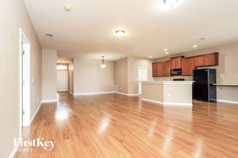 A spacious living room with wooden floors and a kitchen area in the background.