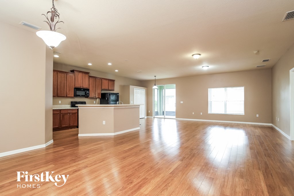 A spacious living room with wooden floors and a kitchen area in the background.