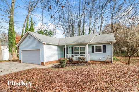 A house with a garage and a front porch is surrounded by trees and has fallen leaves on the ground.