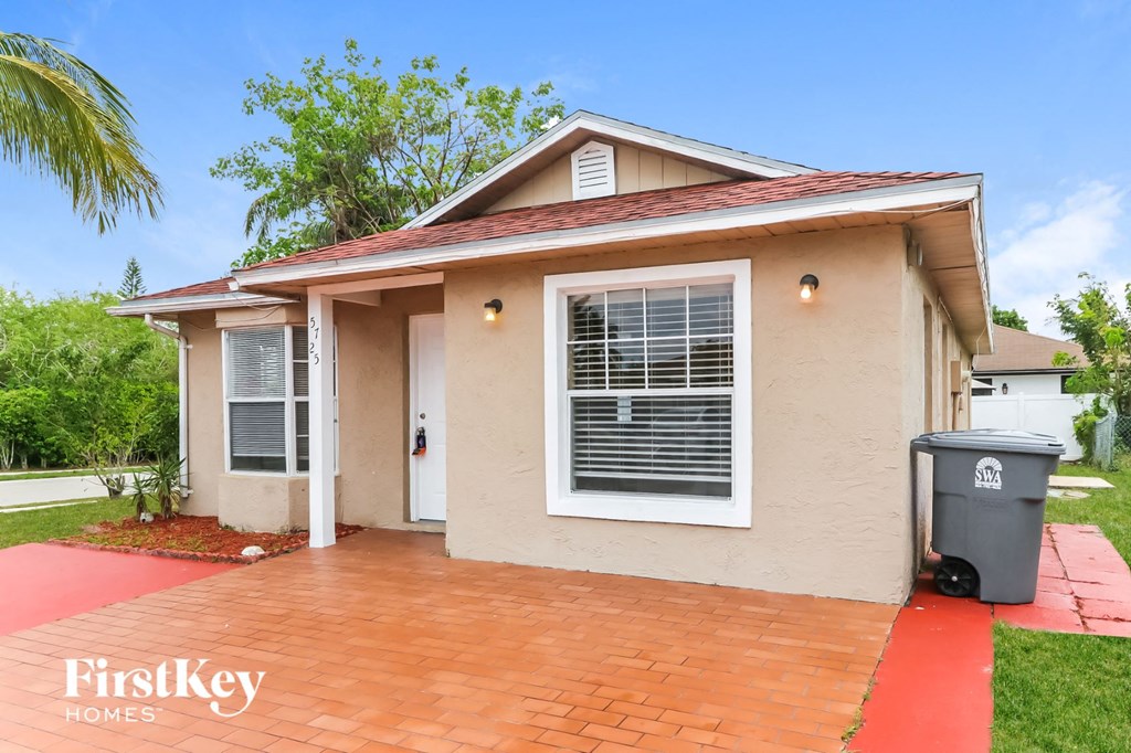 a small tan house with a driveway and a trash can