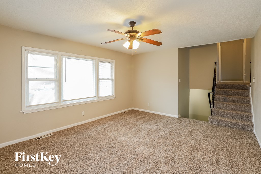 A room with a carpeted floor, a ceiling fan, and a staircase.