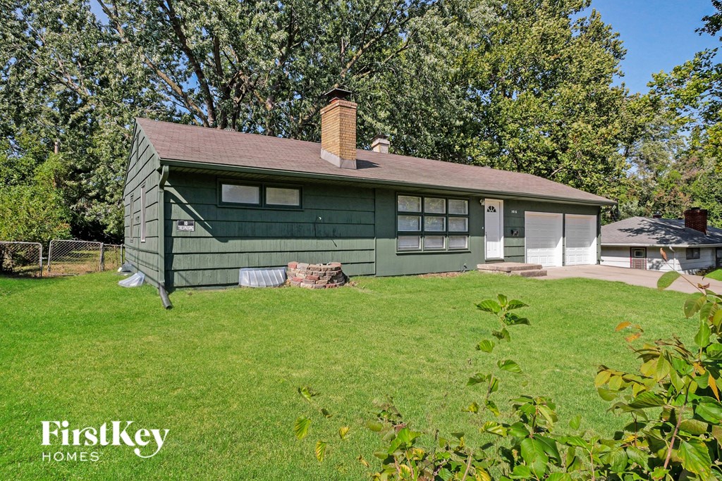 A green house with a brown roof and a chimney is surrounded by a grassy lawn and trees.