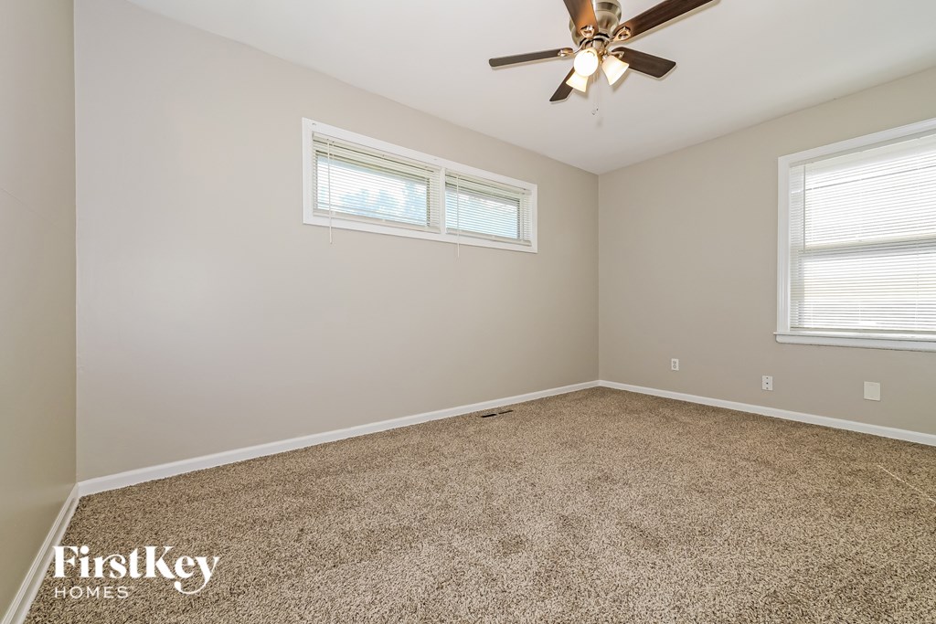 A room with a carpeted floor, a ceiling fan, and two windows.