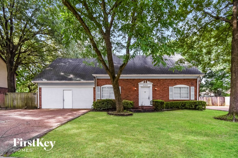 A house with a brick facade and a white door is surrounded by a green lawn and trees.