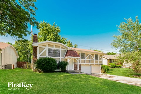 A house with a white and beige exterior is surrounded by greenery and has a clear blue sky above.