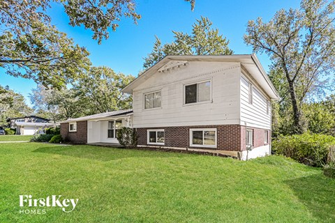 A house with a white exterior and a brick chimney is surrounded by a green lawn and trees.