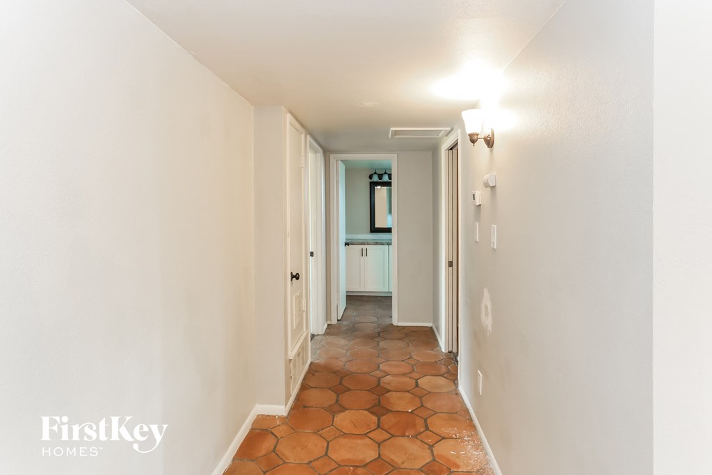 a hallway with white walls and tile flooring and a bathroom in the background