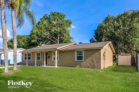 A house with a brown roof and a beige wall is surrounded by a green lawn and trees.