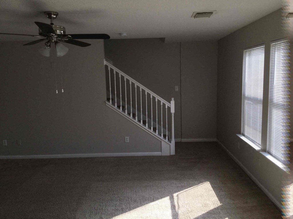 A room with a carpeted floor, a ceiling fan, and a staircase with white railings.