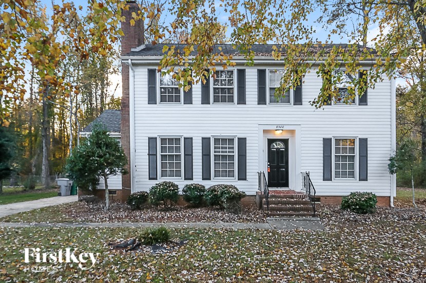A white house with a black door and windows is surrounded by trees with yellow leaves.