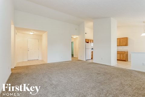 A spacious, empty room with a carpeted floor and white walls, with a door on the left and a kitchenette on the right.