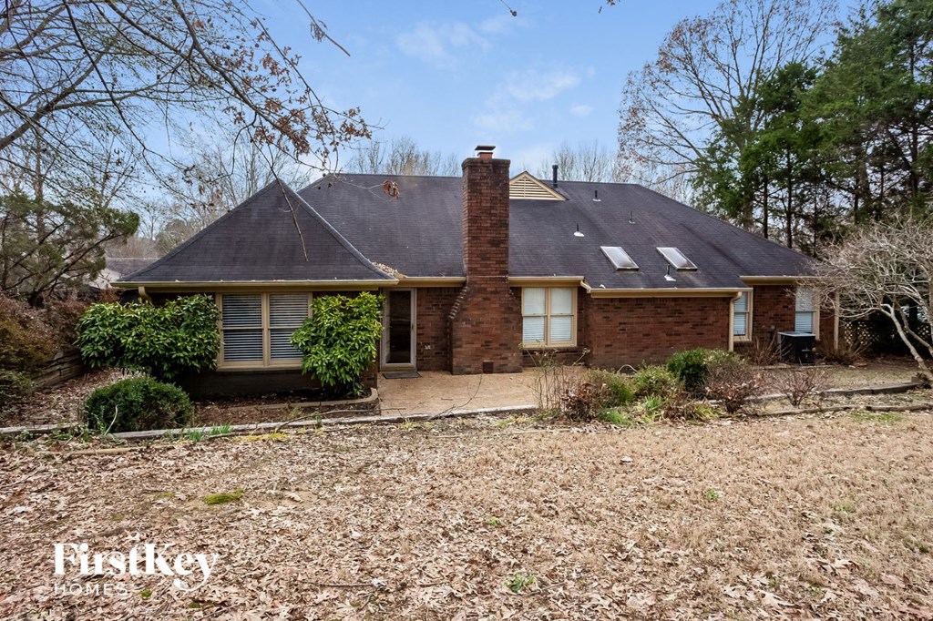 A brick house with a black roof and a chimney is surrounded by trees and bushes.